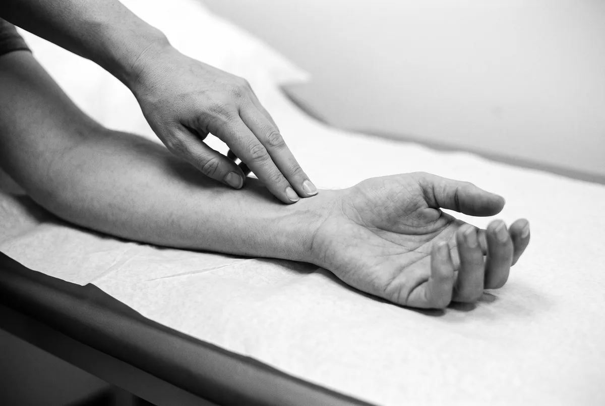 Close-up of a clinician pressing two fingertips against a patient's wrist on an exam table.